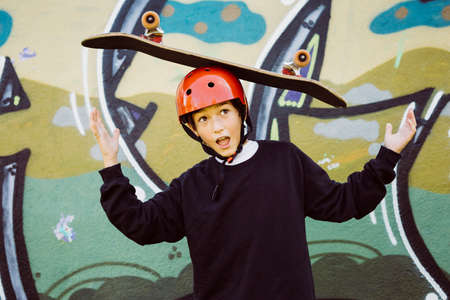 Portrait of a young skater boy wearing red helmet and playing with an old skateboard, in front of a...
