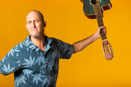 Portrait of an aggressive Musician trying to break his acoustic guitar against a yellow background
