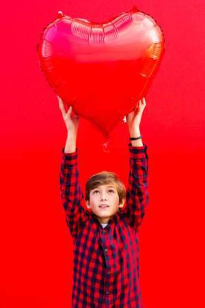 Boy holding a big red heart shaped balloon against a red wallの写真素材