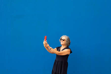 Senior gray haired woman taking a selfie with a red smartphone against a blue wallの写真素材