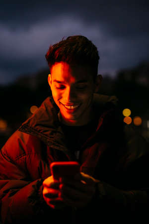 Portrait of a young man on the street with a black coat and using a smartphone, under a red lightの写真素材