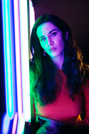 Portrait of brunette caucasian young woman with long hair, standing next to a colored neon lights