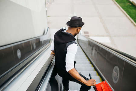 Rear view of a Black young male going down an escalator with a red suitcaseの写真素材