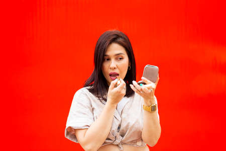 Young brunette woman using lipstick and a small mirror against a red backgroundの写真素材
