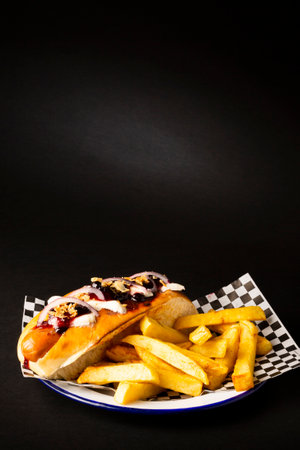Frankfurt sausage with onion, cheese accompanied with french fries on a white plate over a black...
