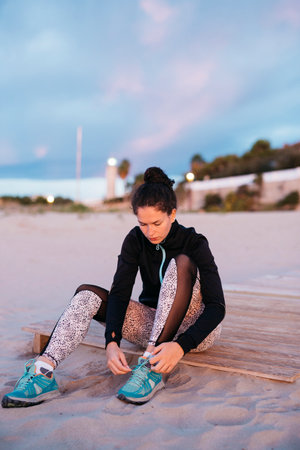 Young woman in sportswear tying laces before working out at the beach at sunsetの写真素材