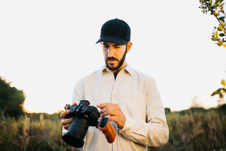 Portrait of a young bearded man, wearing a black cap, with his digital camera at countrysideの写真素材