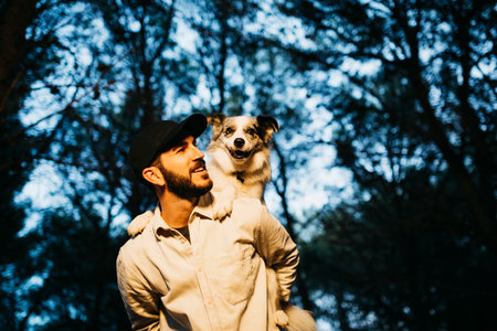 Portrait of a young bearded man, wearing a black cap, carrying a dog on his back at countrysideの写真素材