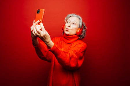 Senior woman wearing red clothes, taking a selfie with red headphones and red smartphone, over a red...