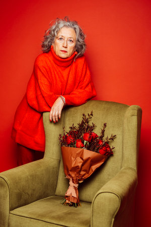 Senior woman wearing red clothes, standing next to a green armchair over a red backgroundの写真素材