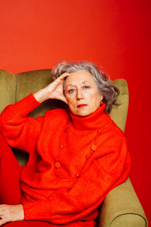 Senior woman wearing red clothes and sitting on a green armchair over a red backgroundの写真素材