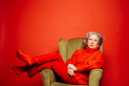 Senior woman wearing red clothes and sitting on a green armchair over a red backgroundの写真素材