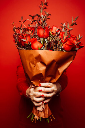 Senior woman wearing red clothes, hiding her face with a red roses bouquet, over a red backgroundの写真素材