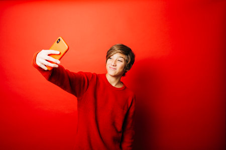 Pre-adolescent boy wearing a red sweater over a red background, taking a selfie with a red smartphoneの写真素材