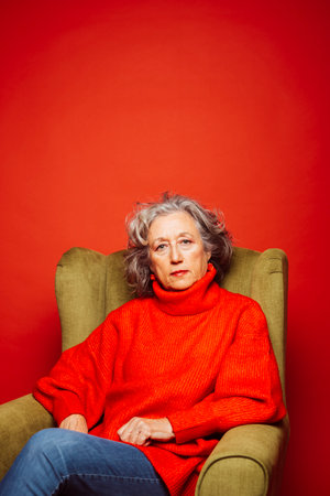 Senior woman wearing red clothes, sitting on a green armchair, over a red background