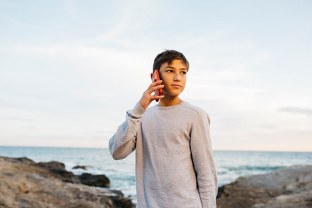 Boy talking on a red cellphone on a rock breakwater, by the seaの写真素材