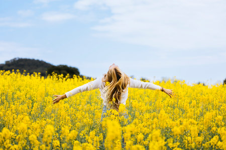 Beautiful young blonde woman with open arms looking at the sky poses amidst a field of blooming yellow rapeseed flowersの写真素材