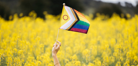 Woman's hand shaking an Intersex-Inclusive Progress Pride Flag amidst a field of blooming yellow rapeseed flowersの写真素材