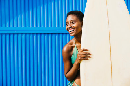Portrait of a surfer standing, in front of a blue background in swimwear, with his surfboard by his side. Happy woman doing beach activities during summer vacation.の写真素材