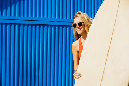 Portrait of a surfer standing, in front of a blue background in swimwear, with his surfboard by his side. Happy woman doing beach activities during summer vacation.の写真素材