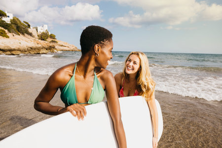 Two smiling women in swimwear carrying a surfboard at the beach during a fun vacation by the sea.の写真素材