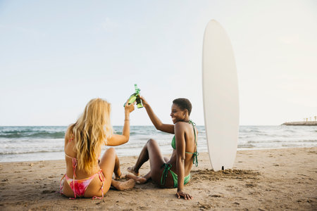 Two cheerful female friends making a toast after a surfing day at the beach. Happy women in bikini with a beer bottle on the shore.の写真素材