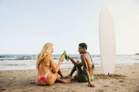 Two cheerful female friends making a toast after a surfing day at the beach. Happy women in bikini with a beer bottle on the shore.の写真素材