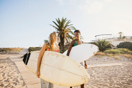Two smiling women in swimwear carrying a surfboard at the beach during a fun vacation by the sea.の写真素材