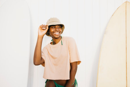 Portrait of a cheerful young black woman standing on a white wall with their surfboards after a day off at the beach.の写真素材