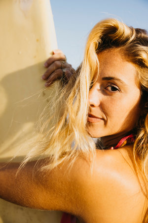 Portrait of a surfer standing on the beach in swimwear, with his surfboard by his side. Happy woman...