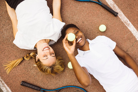 Portrait of two playful young beautiful women with tennis clothes and rackets laying on a tennis court. Two friends sharing a morning of sportive activity playing a tennis match.の写真素材