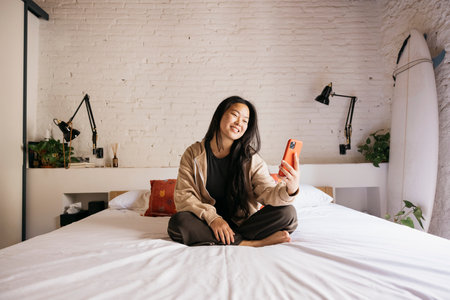 Smiling young beautiful woman sitting on bed and looking at red phoneの写真素材