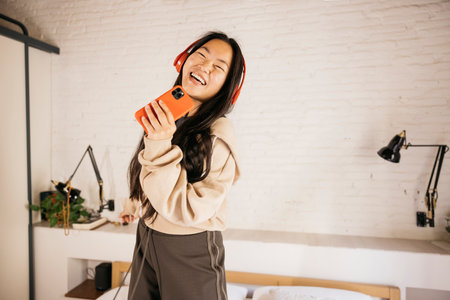 Young chinese woman listening music with headphones and using a phone as a microphone and dancing on a bed.の写真素材