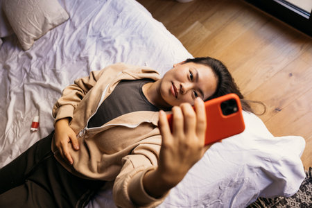 Smiling young beautiful woman lying on bed and using a red phoneの写真素材