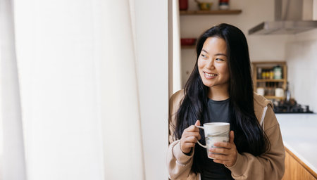 Young beautiful woman with a mug drinking coffee next to a window in a kitchen. Smiling woman enjoying a coffee break at home.の写真素材