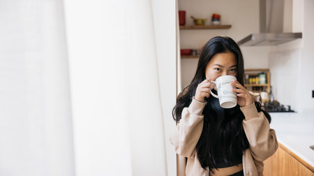 Young beautiful woman with a mug drinking coffee next to a window in a kitchen. Smiling woman enjoying a coffee break at home.の写真素材