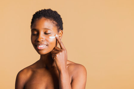 Young black smiling woman with eyes closed applying moisturizing cream on her face. Young woman treating her facial skin with a beauty product.の写真素材