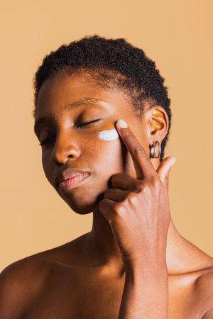 Young black woman with eyes closed applying moisturizing cream on her face. Young woman treating her facial skin with a beauty product.の写真素材