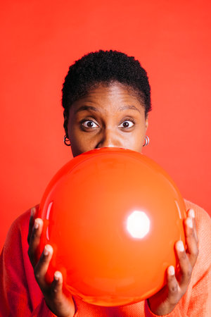Happy young woman blowing a red balloon in front of red background in a studio. Woman with short...