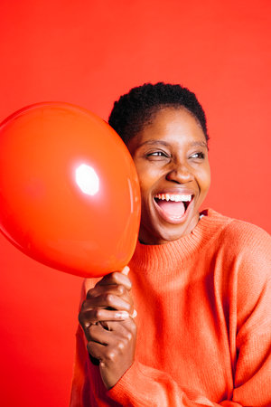Happy young woman blowing a red balloon in front of red background in a studio. Woman with short hair inflating a red balloon.の写真素材