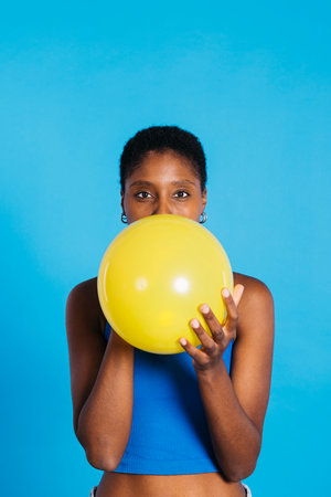 Young woman blowing a yellow balloon in front of blue background in a studio. Woman with short hair inflating a yellow balloon.の写真素材