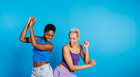 Two young cheerful women dancing in a studio with a blue background.の写真素材