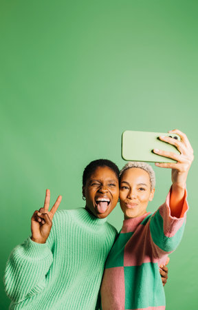 Two young happy women taking a selfie in front of a green background in a studio. Women taking a selfie in a studio.の写真素材