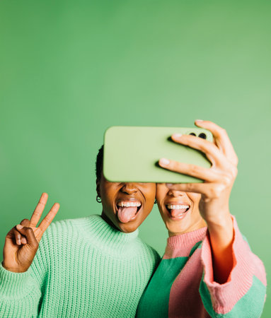 Two young happy women taking a selfie in front of a green background in a studio. Women taking a selfie in a studio.の写真素材