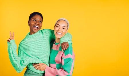 Two young cheerful women wearing green clothes embracing in a studio with a yellow background.の写真素材