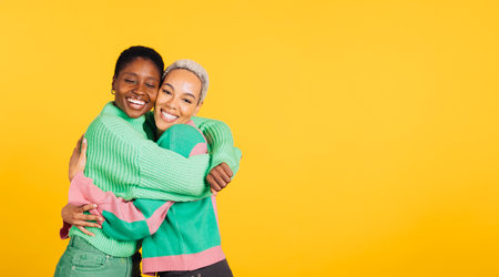Two young cheerful women wearing green clothes embracing in a studio with a yellow background.の写真素材