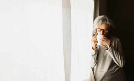 Senior woman standing by a window and drinking coffee on a sunny day at home.の写真素材