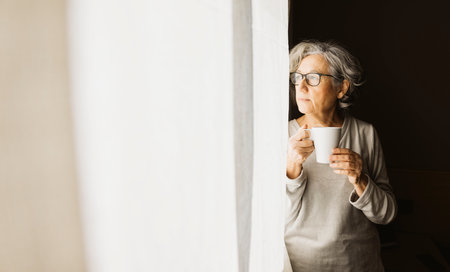 Thoughtful senior woman standing by a window and drinking coffee on a sunny day at home.の写真素材
