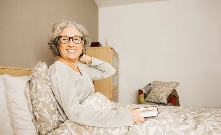 Portrait of a senior woman sitting on bed wearing pajama and reading a book in the morning.の写真素材
