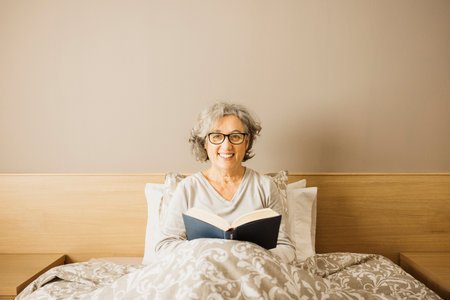 Portrait of a senior woman sitting on bed wearing pajama and reading a book in the morning.の写真素材